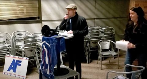Vincent James Foxx, wearing a hat and trenchcoat gives a speech on a megaphone on a makeshift podium. An America First flag drapes the podium, and a DIY America First sign sits to the side along a row of stacked chairs. ASU-CRU’s Julie Hoffman stands behind Foxx, about to hand him a piece of paper