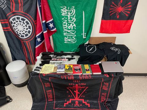 A table with extremist flags draped behind it, guns on the table and books and shirts next to them as well as a handwritten note whose contents are illegible