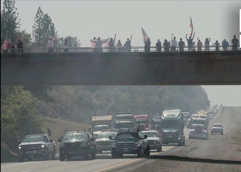Convoy supporters stand on an overpass, watching as the people’s convoy blocks all 3 lanes of a highway. A haze hangs in the air, caused by burning rubber from cars doing burnouts on the roadway.