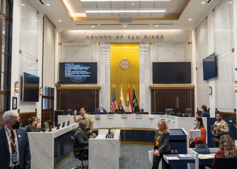 A wide shot of the Board of County Supervisors chamber. On the far wall one can see large monitors, a gold streak up the middle of the wall, flags at the base of the wall, the county seal, and the text "county of san diego" near the ceiling. The Supervisors' seats are emptying in this photo, for a recess. Sheriff's deputies and county workers are moving throughout the frame. It's well lit, and the walls and ceiling are predominantly a white, fine stone.