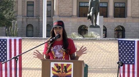 Lindsay Graham standing at a brown podium with an Arizona-themed sign reading “2A rally.” The heavily tattooed woman with long brown hair is wearing a black hat reading “lioness” in red font, her shirt is also red with a bald eagle reading “american woman.” Both her hands are raised as she talks into the extended microphone. Behind her, two American flags are attached to the metal fence in front of the Phoenix Capitol.