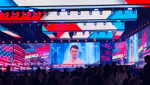 Two large video screens show Charlie Kirk advertising a lunch date with him if you 'text CHARLIE to 71766 to get involved with TPUSA and enter a raffle to have lunch with Charlie Kirk.' The stage lights are red, white and blue, the backdrop has advertisements for AmericaFest and TPUSA