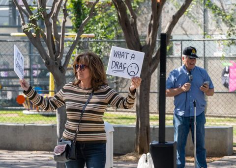 A protester stands in front of a loudspeaker and a mic stand. The protester is holding a sign that reads,"what a drag." The protester in the background is holding a microphone, and reading from a phone.
