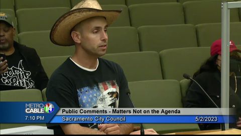 Messano at the public meeting speaking at a podium wearing a straw hat and a shirt with an eagle and a flag on it