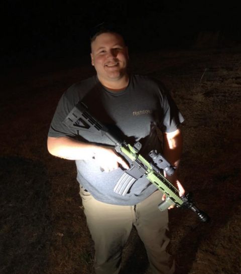 Josh Barham posing with his army green AR-15 rifle standing somewhere in the dark as a flashlight illuminates him.