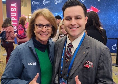 Wendy Rogers and Ryan Sanchez posing for the camera, both giving a thumbs up and smiling. Rogers is wearing a blue jacket that reads “Rogers.” Sanchez is in a grey suit. An AmericaFest badge hangs around his neck. They are both standing in the Media Row section of AmericaFest, a large TPUSA pillar sign and “Turning Point USA AmericaFest” photo wall are behind them.