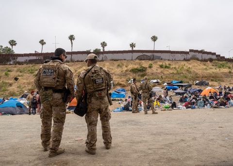 two border patrol agents stand with their backs to the camera looking at the asylum camp