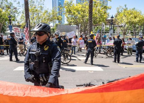 An officer stands beyond the flag. Behind him is a police line, and further beyond is a crowd of fascists.