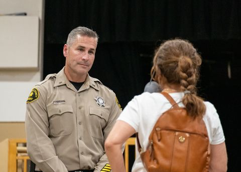 Lt. Wray is seen making eye contact with a woman in a white shirt and brown leather backpack. The woman is shorter than Wray, so he is looking down. The woman is shot from behind, and is out of focus.