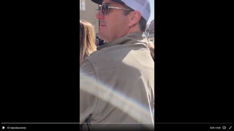 Screenshot of a man in a tan coat, baseball cap and sunglasses