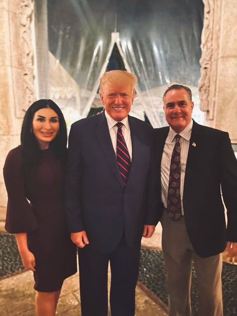Laura Loomer standing to the left of Trump in the center with Guardiola to the right. They're standing in the lobby of Mar-a-Lago between two marble columns with a plastic sheet draped between the columns.