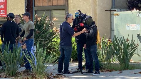 Tony Moon adjusts the body camera on a masked man's chest harness as others stand around outside a building