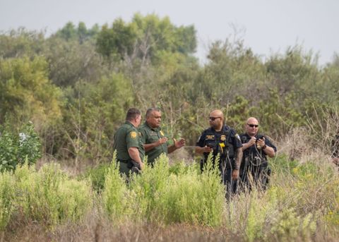 The two agents and two officers are conversing while gesticulating. The two agents are to the left, the two officers are to the right. The foreground is obscured by brush.