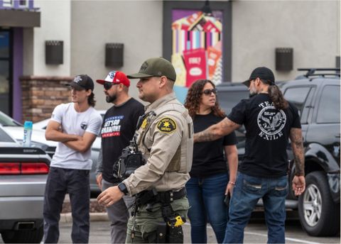 a group of surly men, one in a let's go brandon shirt, stand behind a sheriff's deputy