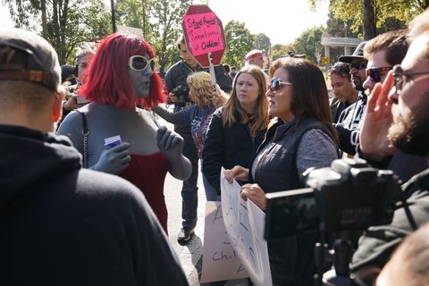 A color photograph of a conversation happening in the middle of a large crowd. The main participants include Emily on the left. She's wearing a grey bodysuit with red dress and red wig. Her face has been painted in the same shade and her glasses are lighter in color. Across from her is Julie, in a grey shirt and slightly darker vest holding a sign. In-between the two but looking towards Emily is Audra Price.