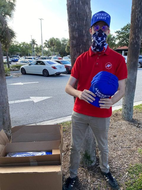A young man in a blue 'America First' baseball cap with an american flag bandana, red polo shirt with an 'AF' logo on it holds a stacks of blue 'America First' baseball caps in front of a box of such caps in a parking lot. He stands next to a tree.