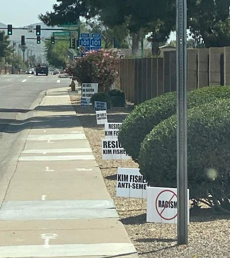 along the road alongside the Deer Valley Unified district office signs placed in the gravel line the sidewalk. The printed signs read 'Kim Fisher anti-semite,' 'Resign Kim Fisher,' and 'racism' crossed out in red
