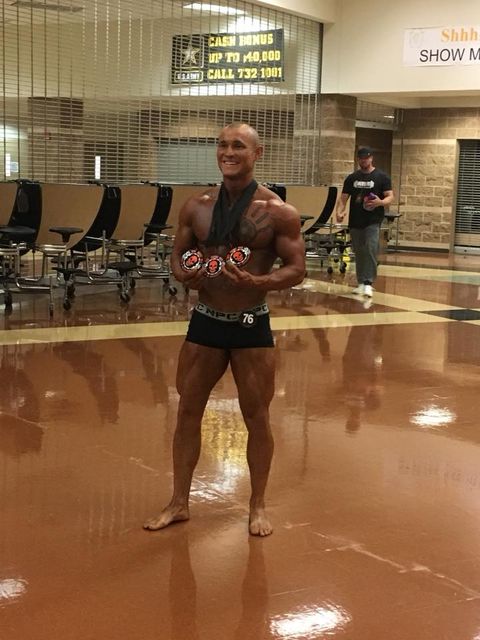 A heavily muscled, almost nude man stands smiling broadly in an empty gymnasium, proudly displaying three competitive medals hanging on ribbons around his neck. His tanned and sculpted body features a large chest tattoo of a hand.