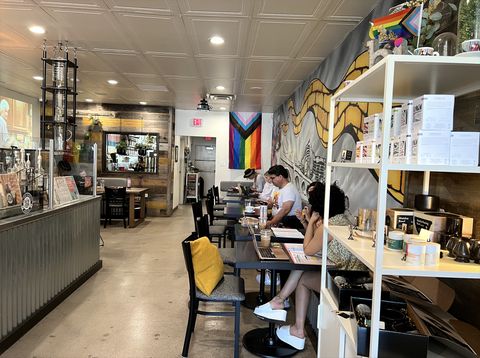David Boyles and several other customers sit inside Brick Road Coffee. One wall is painted with a yellow brick road. Another wall has a trans-inclusive pride flag on display. Golden Girls is being projected on another screen. Items for sale can be seen to the side