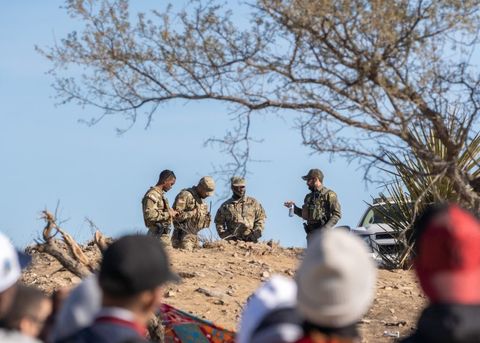 A Border Patrol agent converses with National Guard members on December 14th, 2023. Three men are visible from the waist up, they are wearing camouflage fatigues and standing casually. To their right, a border patrol agent is standing in an olive-green uniform. In the foreground, the tops of asylum-seekers heads are in frame but out of focus.