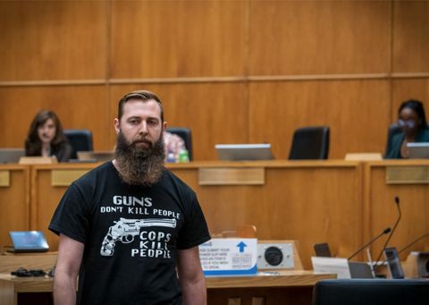  A commenter walks away from the lectern at a PAB meeting on April 27th, 2023. The commenter is facing the camera. They are wearing a shirt that reads, "guns don't kill people, cops kill people" with a graphic of a revolver. Behind the commenter is the Privacy Advisory Board, its members out of focus.