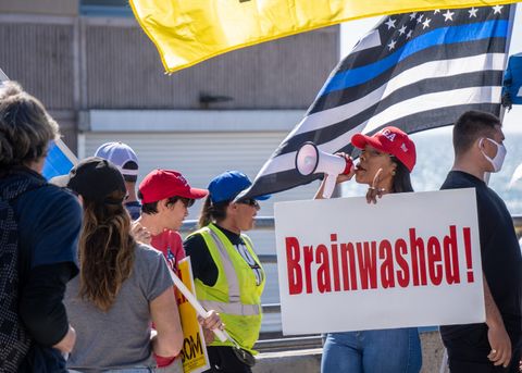 there is a dark skinned maskless woman wearing a maga hat and holding up a white sign in one hand with red text that reads 'Brainwashed!' and the other hand holds a small megaphone at the end. there is a thin blue line flag held by a man wearing a white mask there is the edge of a yellow flag above the thin blue line flag to the left of those two people are several maskless people, some looking towards the person holding th megaphone, others facing away