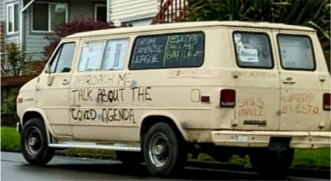 A rickety white van with 'ask me about the covid agenda' spray painted on the door