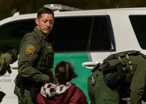 a border agent looks at a young child who's facing him, her back turned to the camera