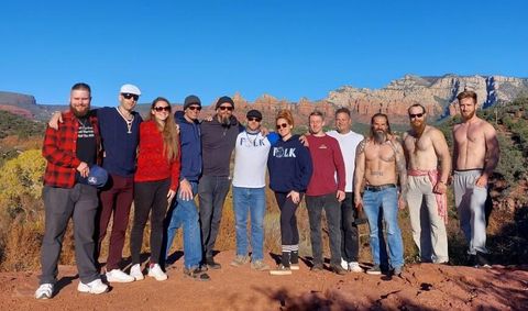 A dozen white people linked arm and arm and posing for a photo in the Arizona desert. Drago is the second from the left and wearing a white golf hat, sunglasses, a blue knitted sweater and burgundy pants and white shoes. Krogstad is next to him wearing a red sweater, black pants, white shoes and sunglasses. In the middle is Sam Ramsey and a woman wearing Asatru Folk Assembly-branded shirts. Three men towards the right end of the shot are shirtless and one is holding a hammer with a visible Sonnenrad tattoo on his shoulder.