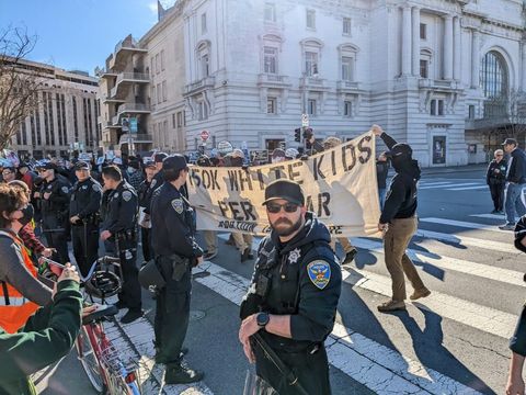 Nazis with their faces covered hold up a banner that says "50k white kids per year abortion is genocide." they're standing behind a police line as counter-protesters make a barrier with bikes between the nazis and cops and themselves