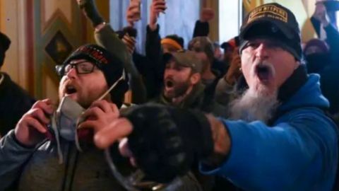 two men shout at the front of a crod inside the capitol building, one on the right in a blue hoodie and oath keepers hat pointing