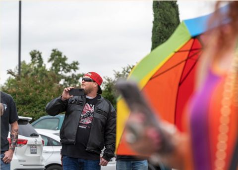 A man in a motorcycle jacket and an America t-shirt films Pride attendees.
