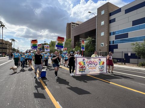 David Boyles and three others hold up a banner for Drag Story Hour Arizona while marching down the street of the Phoenix Pride parade. Others around them hold up colorful signs reading 'We stand with drag story hour.' A group of people wearing blue with colorful rainbow signs follows behind. The sky is blue with fluffy white clouds.