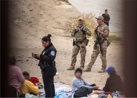 two border patrol agents stand around in the back, one with a rifle, as a homeland security official in a blue uniform looks at a notepad in the foreground as she stands in the middle of a group of asylum seekers