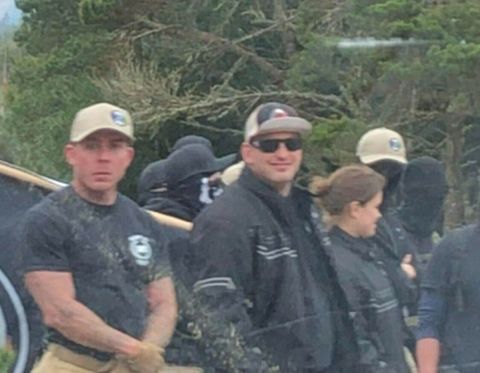 A neo-Nazi at left with his face uncovered wearing a tan hat looks into the camera. He's flexing slightly less hard as before and doesn't look as jaw clenched. To his left is a smiling Dustin Grant.

