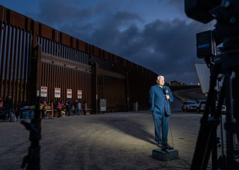 a newsman stands in front of the border fence in the dark with a microphone