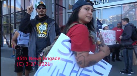 Screenshot of a video of Ed Badalian, wearing sunglasses, a baseball cap and a shirt with a lion on it behind Kennedy Lindsey, wearing a backwards baseball cap and holding a 'save the children' sign.