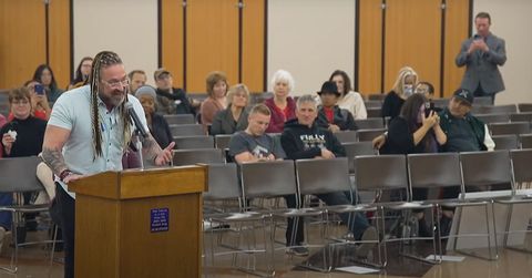 Louis Uridel, a white man with dreadlocks, stands at a podium and speaks to the Oceanside Unified School District board of education, who is off-screen. There are several people behind him, out of focus. One of those people is standing—Bryce Henson, filming Uridel and holding his phone with both hands.