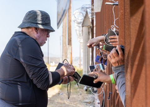 an activist charges phones for people who are sticking their hands through the border fence to hand them to him