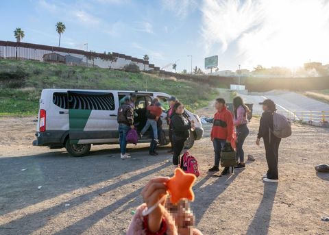 In the middle of the frame, people are exiting a Border Patrol van. They carry personal belongings and children. The sun is behind them, and they all cast long shadows against the warm-orange dirt. The sky is blue, but it's nearly evening. In the foreground, out of focus, a small child is smiling and raising her hand to the camera. In her hand is a small orange star-shaped plushie.