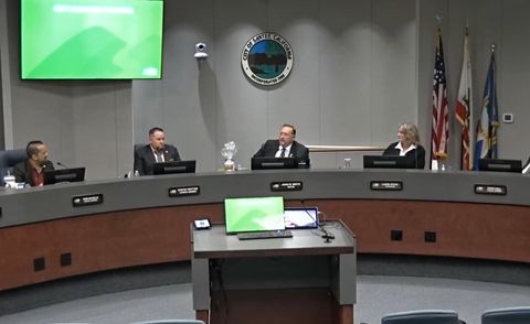 A photo of the live-feed from Santee city council. Four council members are visible behind the dais, including mayor John Minto. The crest of the city of Santee is visible above Minto's head. The podium used for public comment is unoccupied.