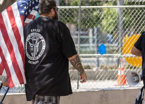 Parmer is standing in a black shirt with plaid shorts. He has short brown hair and a beard. He has forearm tattoos, and is carrying an American flag.