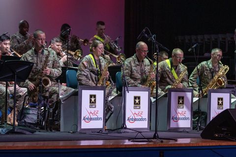 11 band members playing on stage in the Army National Guard Band. The only woman in the group is Ashley Drago/Krogstad in the front row alongside the rest of the camouflage-wearing saxophone players. The podiums in front of the saxophone players is branded with the U.S. Army logo and reads “Jazz Ambassadors.”