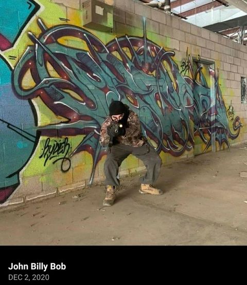 gonzalez in front of some graffiti pointing a rifle at the camera wearing a balaclava