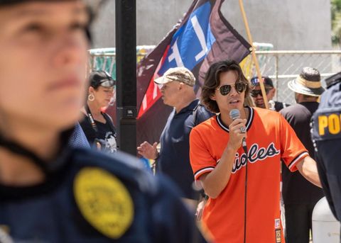 a protester with ear-length brown hair and sunglasses speaks into a microphone. He is in an Orioles jersey, and a LEXIT flag is visible behind him.