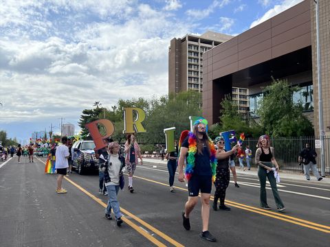 A crowd of people marching in Phoenix Pride walk down the parade street holding up large colorful letters that spell out 'PRIDE.' The person leading the group is wearing a rainbow boa, angel wings, and sunglasses. The sky is blue and filled with fluffy white clouds. Far behind another colorful crowd is carrying even larger colorful letters spelling out 'LOVE.'