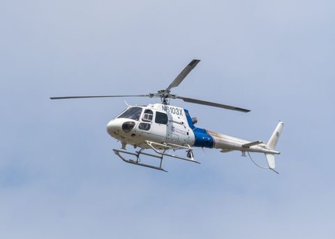 A white helicopter is in the center of the frame, in the sky. It has a blue stripe and a Customs and Border Protection logo.
