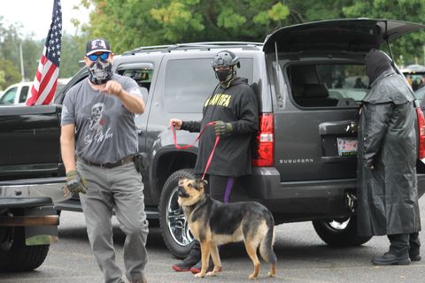 A man points his index finger at the photographer while another man holds a german shepard on a leash in front of some parked cars.
