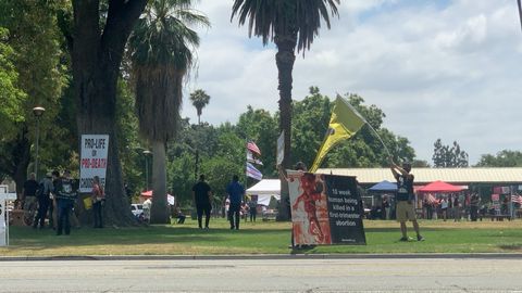 A man waves a yellow flag on the sidewalk of a park next to some anti-abortion signs while people gather inside the park