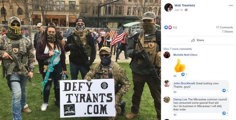 Militiamen standing in camo and gear with long guns clustered around a sign that says defy tyrants dot com. there's a facebook comment from John Brockhoeft that says 'looking great, crew! thanks, guys!'